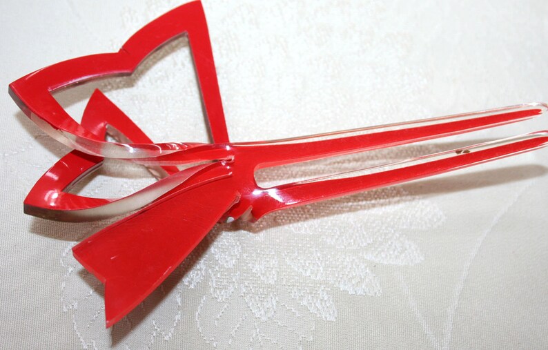 May include: A red plastic hair clip shaped like a bow with two prongs. The bow is translucent and has a glossy finish.