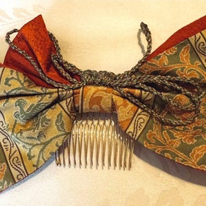 May include: A traditional Japanese hair comb with a large bow made of patterned fabric. The bow is in shades of brown, green, and gold, with a floral design. The comb is silver and has a decorative design.