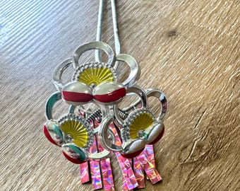 Puede incluir: Tres peines de pelo plateados con diseños de flores rojas, amarillas y blancas. Las flores tienen un centro en forma de abanico y están unidas a cintas iridiscentes de color rosa y plateado.