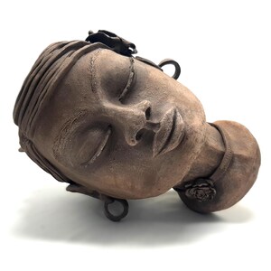 Puede incluir: Una escultura de terracota de una cabeza de mujer con los ojos cerrados, una rosa en el cuello y detalles metálicos. La escultura está inclinada hacia un lado.