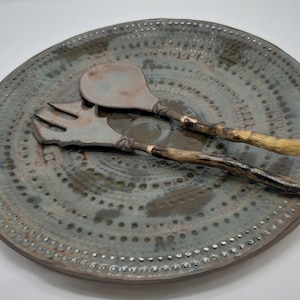 May include: A ceramic serving plate with a blue and brown glaze and a repeating pattern of small circles. The plate has a set of ceramic serving utensils with wooden handles.