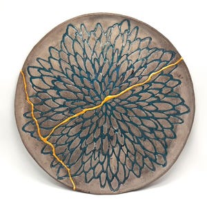 May include: A round ceramic plate with a blue and gray floral design and gold cracks. The plate is a decorative piece with a unique design.