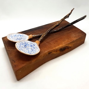 Puede incluir: Dos cucharas de servir con cuencos de cerámica moteada azul y blanca y mangos de madera natural descansan sobre una tabla de cortar de madera oscura. Las cucharas tienen un diseño rústico único, ideal para servir ensaladas.