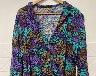 Könnte beinhalten: Ein Vintage-Hemd mit Knöpfen und einem lebendigen, mehrfarbigen Blumen- und Tiermuster. Das langärmlige Hemd hat einen V-Ausschnitt und goldfarbene Knöpfe. Die Farbpalette umfasst Türkis, Lila, Schwarz und Beige.