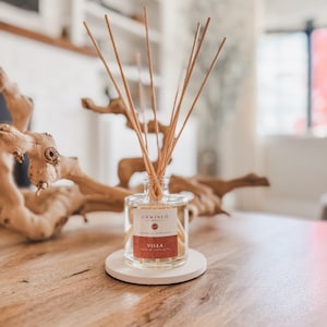 May include: A clear glass reed diffuser bottle with a white label featuring the text UNWINED, Signature Collection, and VILLA in red and black ink. The bottle contains a light-colored liquid and several thin wooden reeds extending upwards.