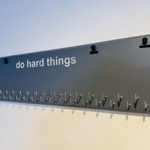 May include: A dark grey wooden board with the text "do hard things" in white lettering. The board has multiple small silver hooks along the bottom edge and two black clips at the top.