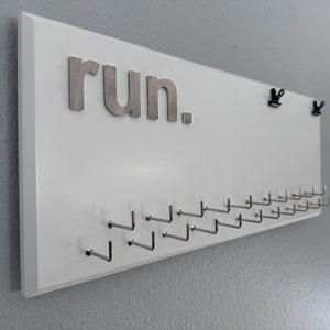 Running Medal Holder white with 3D lettering in aluminium or acrylic. White medal hanger with 3D &quot;run.&quot; wording, 21 hooks and BIB holder.