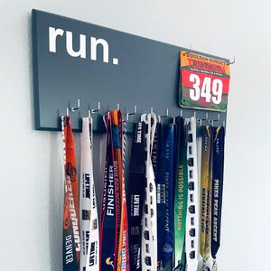 May include: A dark gray rectangular wall-mounted medal hanger featuring the white text "run." and multiple silver hooks. A "Boulder Sunset Triathlon" race bib with the number 349 is attached to the top right, with various colorful athletic ribbons.