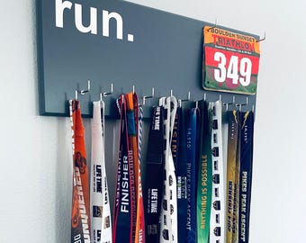Peut inclure: Un porte-médailles mural rectangulaire gris foncé avec le texte blanc "run." et plusieurs crochets argentés. Un dossard de triathlon "Boulder Sunset" portant le numéro 349 est fixé en haut à droite, avec divers rubans sportifs colorés.