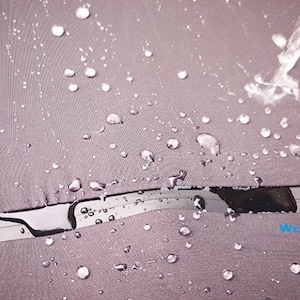 May include: Close-up of a gray fabric umbrella canopy with water droplets on the surface. Water is being poured onto the fabric, demonstrating its waterproof properties. The umbrella has a black and silver metal frame.