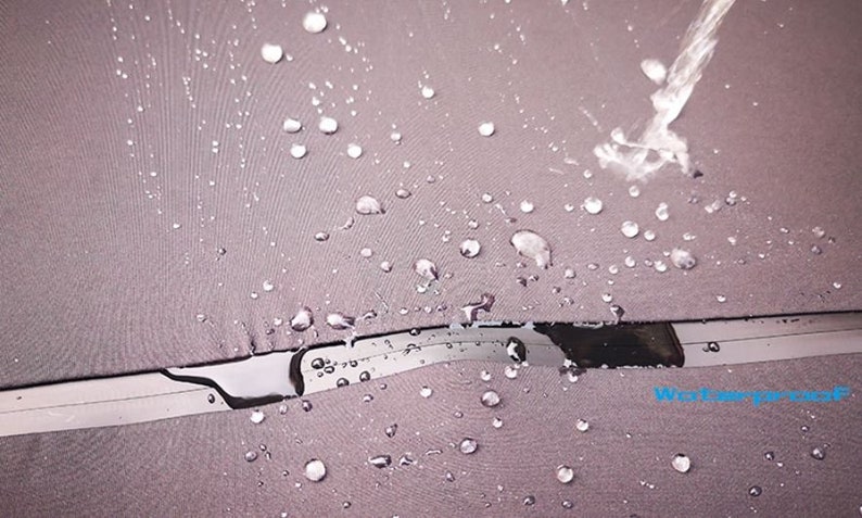 May include: Close-up of a gray fabric umbrella canopy with water droplets on the surface. Water is being poured onto the fabric, demonstrating its waterproof properties. The umbrella has a black and silver metal frame.
