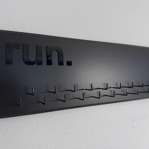 May include: Black medal display with the word "run." and multiple hooks for hanging medals. The rectangular display is mounted on a white wall. A great gift for runners to showcase their achievements.