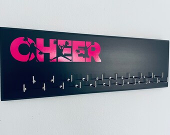 May include: A rectangular black wall-mounted display rack featuring the word CHEER in bright pink lettering. The letters incorporate silhouettes of cheerleaders holding pom-poms. The rack includes two staggered rows of silver metal hooks.