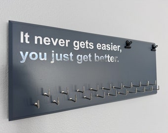 May include: A rectangular dark blue wall-mounted display board featuring the white text It never gets easier, you just get better. The board includes two black metal clips at the top and a series of silver-colored metal hooks arranged in two rows.