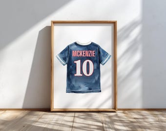 May include: Watercolor print of a blue jersey with the name "MCKENZIE" and the number "10" in red and white. The jersey has white stripes on the sleeves and a white collar. The artwork is framed in a wooden frame.