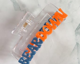 May include: Clear plastic hair claw clip with the words "BEAR DOWN" in blue and orange raised letters. The clip has a gold-coloured spring mechanism and is set against a white and grey marbled background.