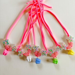 May include: A collection of bright pink elastic hair ties, each adorned with a translucent star charm filled with multicolored confetti, a small pink heart-shaped bead, and a single tooth-shaped charm in white, blue, green, or yellow.