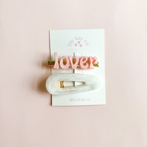 May include: A set of two hair clips. One clip is a white, glittery, oval-shaped clip. The other clip is a gold bar with the word "lover" in pink letters.