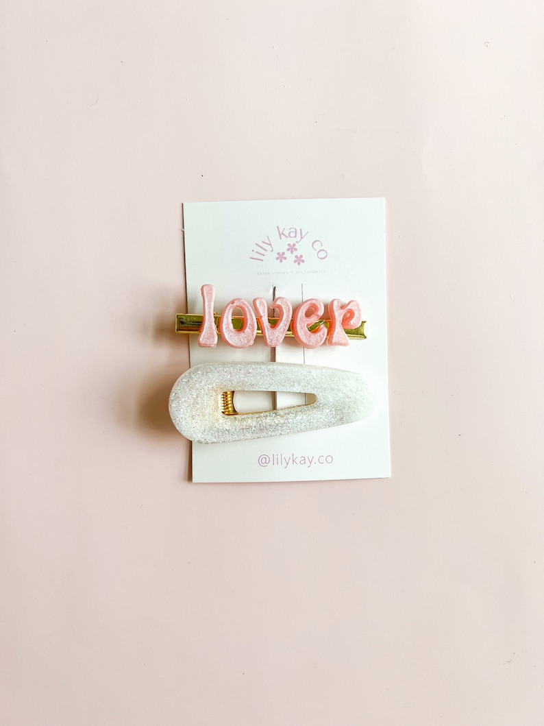 May include: A set of two hair clips. One clip is a white, glittery, oval-shaped clip. The other clip is a gold bar with the word "lover" in pink letters.