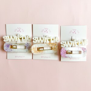 May include: Three white hair clips with the word "Swiftie" written in white letters with gold glitter. Each clip has a different color glitter: purple, gold, and pink.