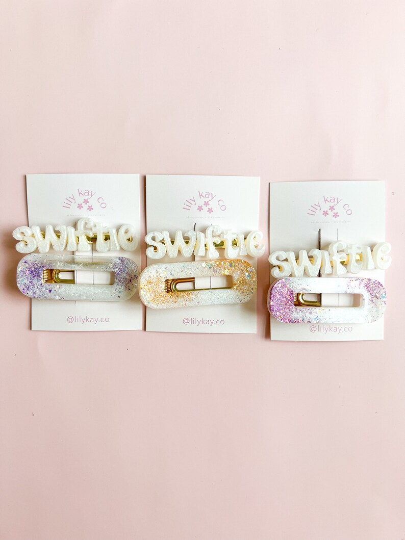 May include: Three white hair clips with the word "Swiftie" written in white letters with gold glitter. Each clip has a different color glitter: purple, gold, and pink.