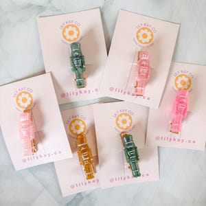Puede incluir: Seis pinzas para el pelo con forma de cascanueces de colores variados, incluyendo verde, rosa y dorado. Cada pinza está unida a una tarjeta pequeña con el texto "LILY KAY CO" y "@lilykay.co".