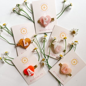 May include: Five heart-shaped hair clips with letters A, L, P, S, and D. Each clip is made of resin and has a gold metal bar. The clips are arranged on a white background with white daisies.