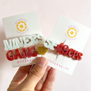 Könnte beinhalten: Zwei Haarklammern mit den Worten "Winners Gang" und "49ers" in roten und weißen Buchstaben. Die Klammern befinden sich auf einem weißen Hintergrund mit einem goldenen Herzen.
