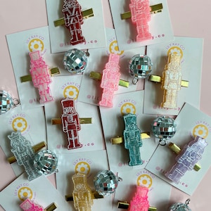 May include: A collection of 10 colorful nutcracker hair clips with glitter and a disco ball charm. The clips are on white cards with the text "Lily Kay Co" and a flower logo.