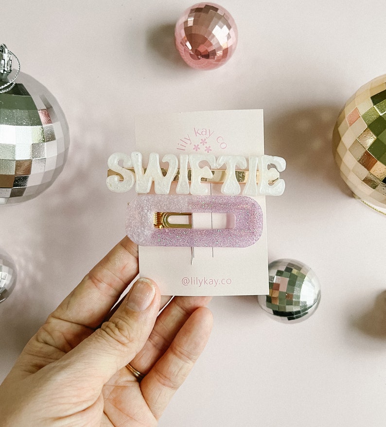 May include: A white glitter hair clip with the word "SWIFTIE" in raised letters. The clip is on a pink background with a small white card that says "lily kay co" and "@lilykay.co".