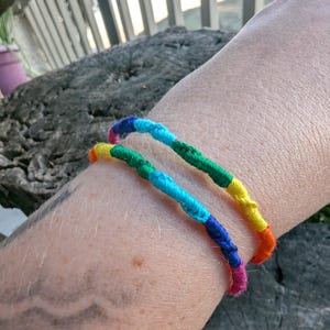 May include: Two rainbow-colored friendship bracelets. Each bracelet is made of tightly wound threads in a spectrum of colors, including red, orange, yellow, green, blue, and purple. The bracelets are worn on a wrist.