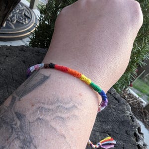 May include: A rainbow-colored friendship bracelet with a braided design. The bracelet features a spectrum of colors, including red, orange, yellow, green, blue, and purple. The bracelet is tied with a tassel at the end.