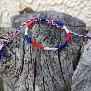 May include: A red, white, and blue friendship bracelet made with braided string. The bracelet is tied in a knot and has a tassel on each end.