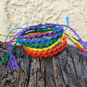 May include: A set of rainbow colored friendship bracelets, woven with a spiral pattern. The bracelets are made of colorful string and are tied together in a knot.