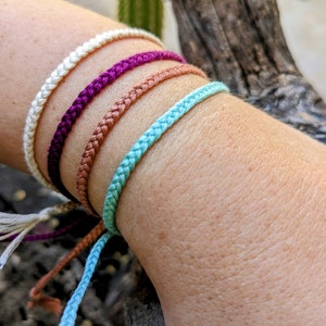 May include: Four braided friendship bracelets in white, purple, brown, and light blue colors. The bracelets are tied together at the ends.