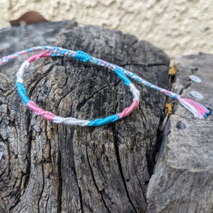 May include: A braided friendship bracelet with pink, blue, and white threads. The bracelet is tied in a knot and is lying on a piece of weathered wood.