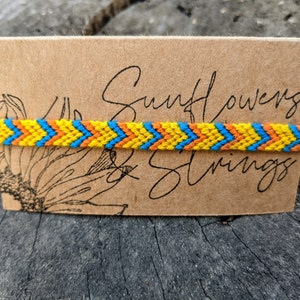 May include: A yellow, orange, and blue friendship bracelet with a chevron pattern. The bracelet is on a brown card with the text "Sunflowers & Strings".