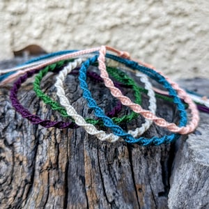 May include: A set of six colorful friendship bracelets made with twisted cord. The bracelets are in shades of green, blue, purple, and pink.