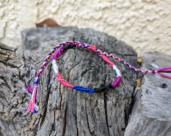 May include: A bi pride flag friendship bracelet. The bracelet is made with pink, purple, blue, black, and silver threads. The bracelet is displayed on a weathered wooden surface.