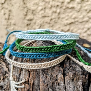 May include: A collection of handmade woven bracelets in various colors, including light blue, green, and cream. Each bracelet features a braided design and is displayed on a piece of weathered wood. The bracelets have tassels at the ends.