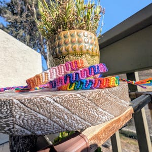 May include: Three colorful friendship bracelets with intricate woven patterns rest on a wooden surface. The bracelets feature a vibrant array of colors, including orange, pink, blue, and yellow. Each bracelet has a braided tassel at the end.
