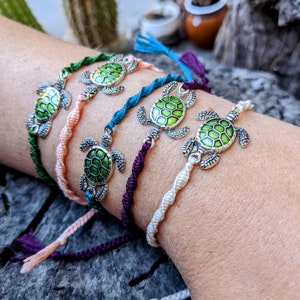 May include: A set of six colorful friendship bracelets with a silver turtle charm on each. The bracelets are made of braided string and come in a variety of colors, including pink, blue, green, and white.