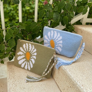 以下が含まれることがあります： デイジーの花柄のクロシェバッグが2つ。左側のバッグは緑色で、白いデイジーと黄色のセンターがあります。右側のバッグは薄い青色で、白いデイジーと黄色のセンターがあります。両方のバッグには白と緑のタッセルが付いています。