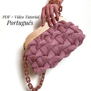 Può includere: Una pochette rosa in maglia con un manico superiore in legno e una tracolla a catena di plastica rosa. La borsa è tenuta da una mano che indossa un braccialetto a catena di plastica rosa. Il testo "PDF + Video Tutorial Português" è visibile nell'immagine.