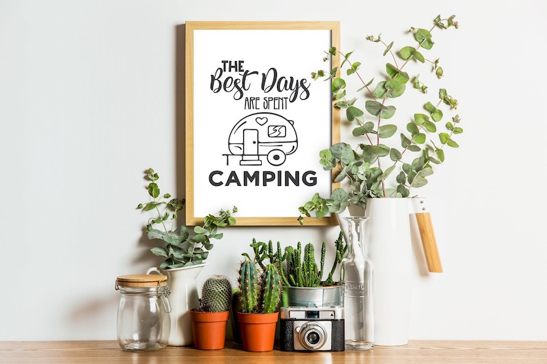 Puede incluir: Una impresi&oacute;n enmarcada con el texto "The Best Days Are Spent Camping" y una ilustraci&oacute;n en blanco y negro de una caravana. La impresi&oacute;n est&aacute; rodeada de plantas en macetas y una jarra blanca.