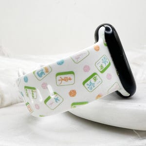 May include: White watch band with a pattern of green, pink, and orange symbols. The band has a black clasp and is designed to fit a smartwatch. The symbols appear to be related to a game.