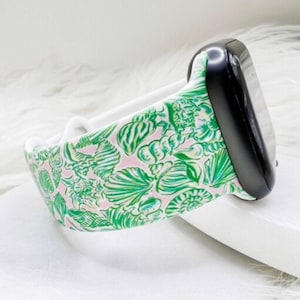 May include: A black smartwatch with a white band and a patterned strap. The strap features a pink background with green and white seashell designs. The watch is displayed on a white surface.