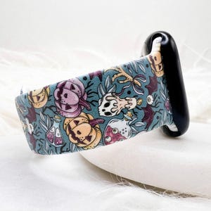 May include: A teal watch band with a Halloween-themed pattern. The design includes pumpkins, skulls, potion bottles, and bats in shades of purple, orange, and white. The watch band is attached to a black watch face.