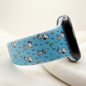 May include: A light blue smartwatch band featuring a repeating pattern of Snoopy characters swimming, musical notes, and yellow flowers. The band has a black frame and is designed for a smartwatch.