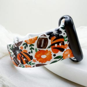 May include: A white silicone watch band featuring a repeating pattern of brown footballs, orange and black tiger-striped shapes, and orange flowers with green leaves. The band is attached to a black watch connector piece on one side.
